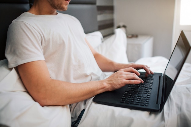 Cut view of young man in bed this morning. He work by holding laptop and typing on keyboard. Work from home. Daylight.