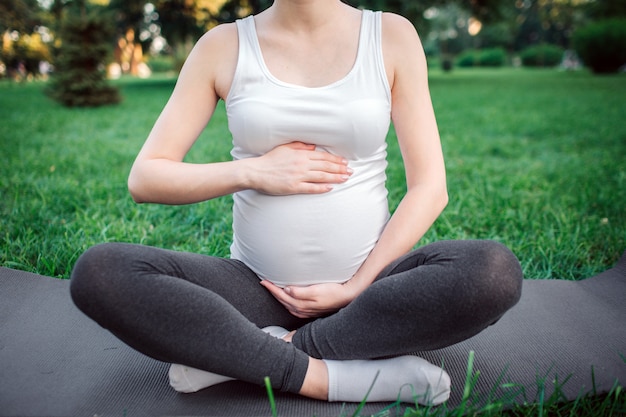 Cut view of young pregnant woman sit on yoga mate in park. She hold hands around belly. Model sit in lotus pose.