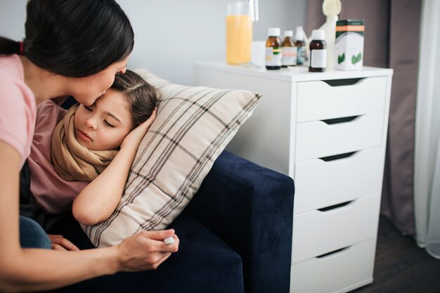 Cut view of young woman kissing forehead of her daughter. She sit besides her couch. Sick girl lying there. Mother hold thermometer in hand. They are in one room.