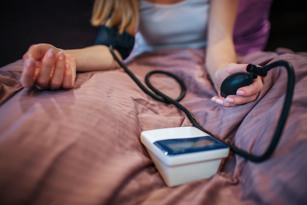 Photo cut view of young woman sitting on bed and mesuring blod pressure with special tool. she squizes pump. woman is concentrated.