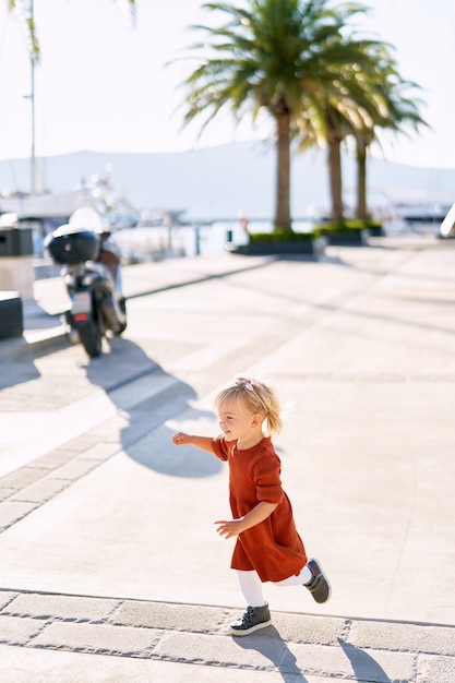 Cute 18-month old girl in a terracotta dress and white tights is running on a boat pier on a sunny day.  