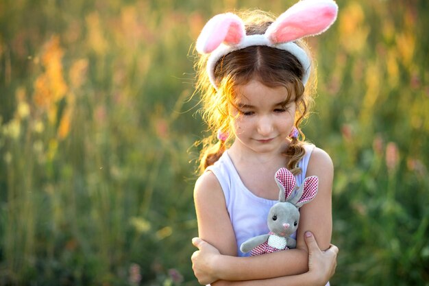 Photo cute 5yearold girl with rabbit ears gently hugs a toy rabbit in nature in a blooming field in summer with golden sunlight easter easter bunny childhood happy child springtime