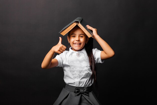 Cute 7 year old schoolgirl with a book on her head, shows the class, isolated on a dark background.