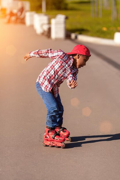 Милый активный афроамериканский мальчик на роликах катается на коньках по асфальтовой дороге на летнем закате Ребенок на роликах учится первым шагам Блики от линз