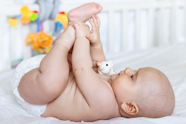 A cute adorable  baby in the diaper  is playing with his legs lying on his back on the bed and looking on his legs