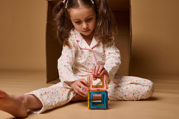 Cute adorable Caucasian baby girl sitting ahead a cardboard box and building a colorful house using square and triangle magnetic construction blocks Fine motor skills development concept