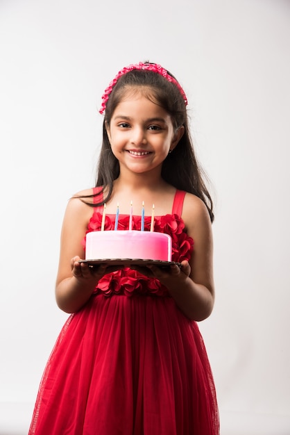 Carina adorabile bambina indiana o asiatica che festeggia il compleanno mentre tiene in mano una torta di fragole e soffia candele a tavola o in piedi isolata su sfondo bianco o rosso