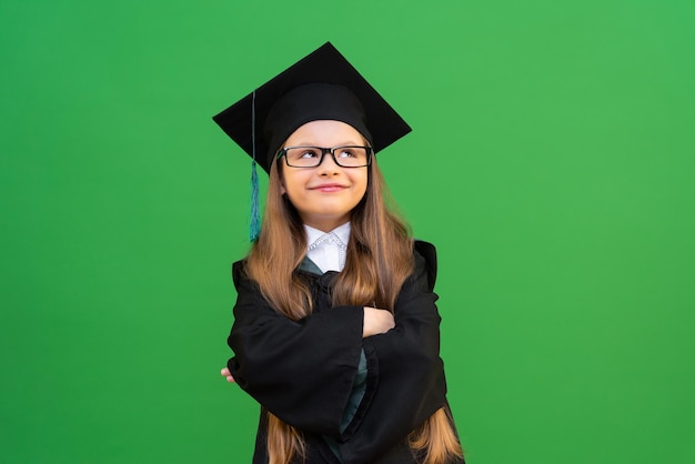 かわいい愛らしい女子高生は学校で勉強するのがとてもうれしいです、修士のガウンと帽子の学生は証明書を待っています、眼鏡をかけた緑の孤立した背景の美しい女の子、