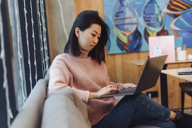 Милая взрослая девушка играет в ноутбуке в кафе. Азиатская женщина, работающая в кафе на ноутбуке. Концепция фрилансера.