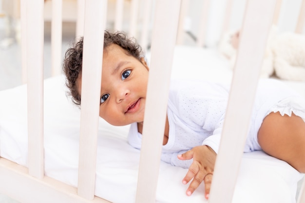 Cute African-American little baby in a white bed for sleeping looks over the side