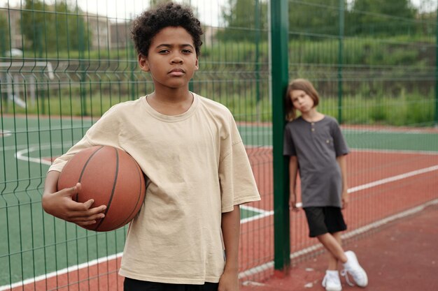 Cute african american schoolboy with ball looking at camera against fence