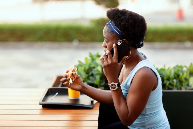 Photo cute african woman having lunch outdoors in city cafe. a girl is talking on the phone during lunch.