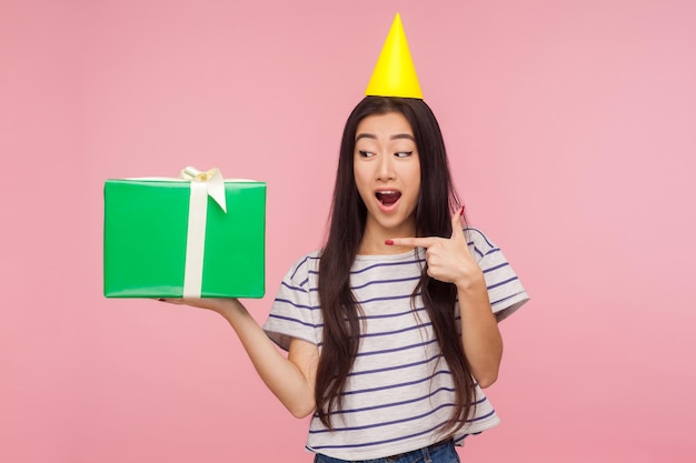 Cute amazed girl with party cone on head pointing at gift box and looking with amazement showing awesome birthday present offering anniversary bonus indoor studio shot isolated on pink background