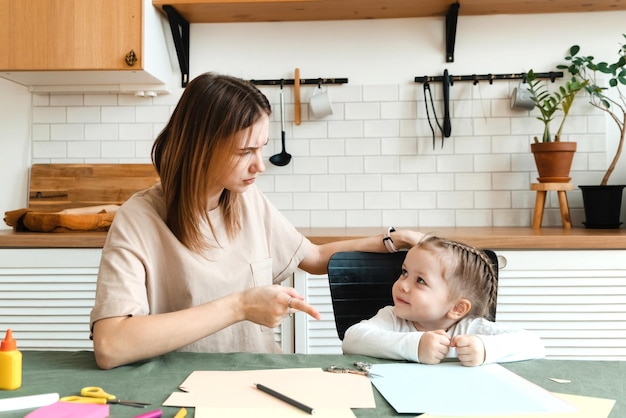 Милая злая маленькая дошкольница делает творческую домашнюю работу со своей мамой, сидя за столом в гостиной и споря о проблемах с изготовлением поделок для детского сада