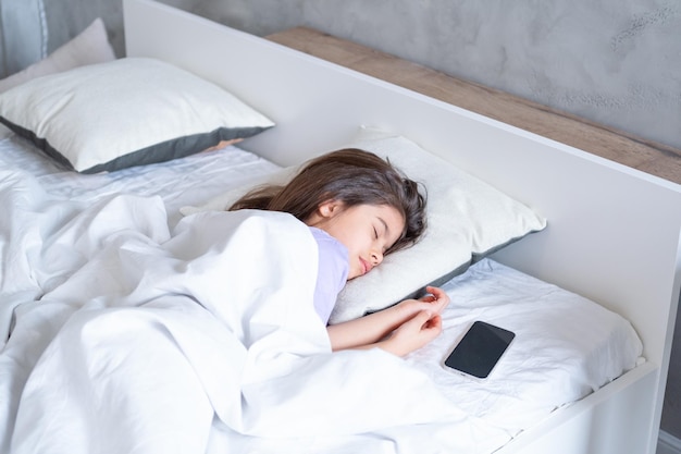 милая арабская девочка с длинными волосами спит на белом постельном белье дома рядом с ней телефон