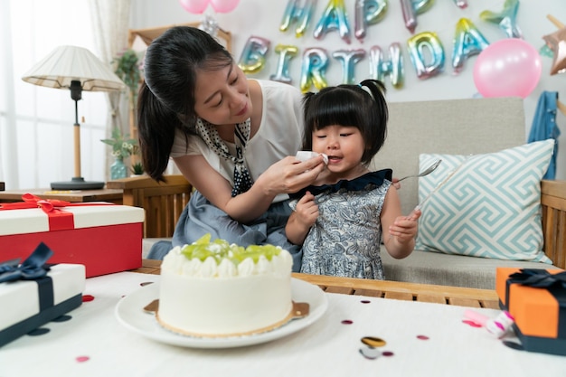 彼女の母親が彼女の口にクリームを拭き取っている間、手にナイフを持ってテーブルの上のおいしいケーキを見ているかわいいアジアの女の赤ちゃん。自宅で楽しむ誕生日のお祝い
