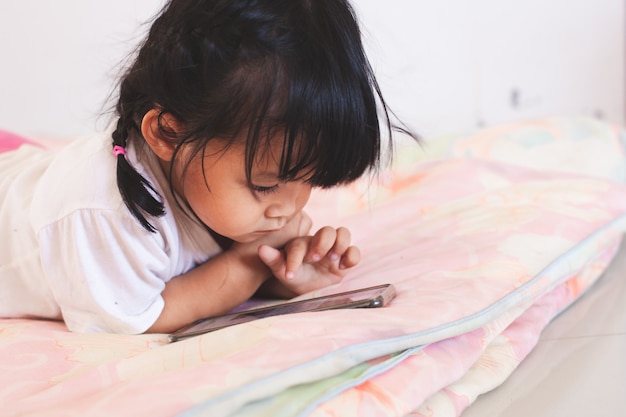 彼女の部屋で彼女のベッドに横になっているスマートフォンを弾くかわいいアジアの赤ちゃん女の子