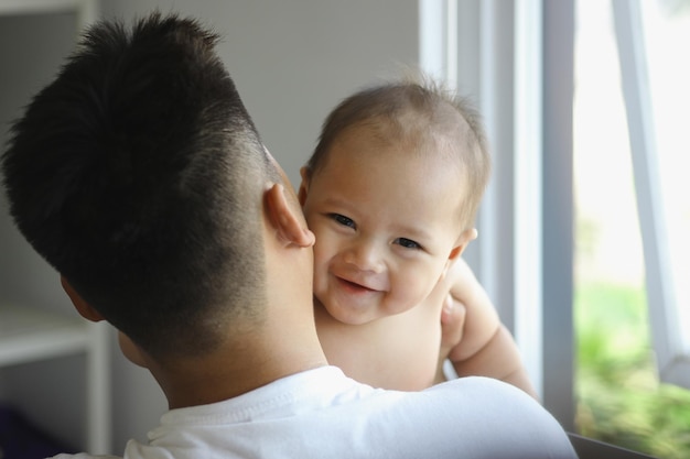 사진 사랑스러운 아시아 아기가 거실의 창문 근처의 아버지의 어깨에 미소 짓고 있습니다.