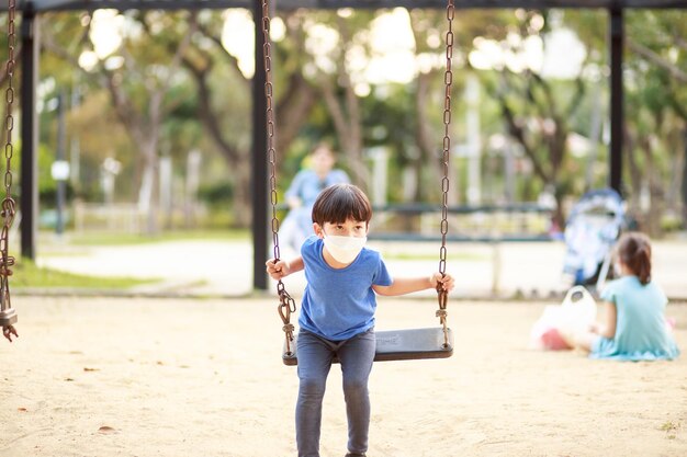 A cute asian boy wearing a mask is playing on a swing in the\
playground during the daytime in summer outdoor activities play\
makes ideas believe external education portrait