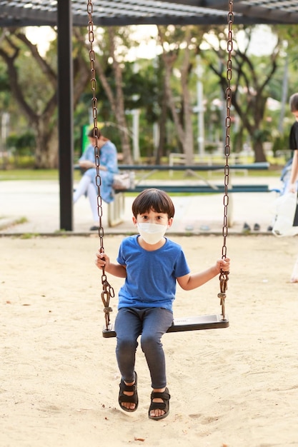 A cute asian boy wearing a mask is playing on a swing in the
playground during the daytime in summer outdoor activities play
makes ideas believe external education portrait