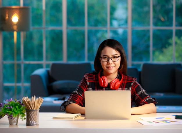 Cute Asian businesswoman working at home living room with twilight sky in outside of house background. She works with laptop notebook computer and looking to camera.