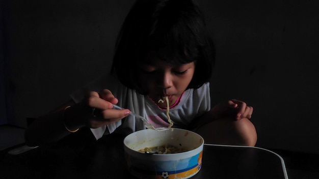 Foto carina bambina asiatica che mangia deliziosi noodles istantanei con la forchetta sullo sfondo scuro