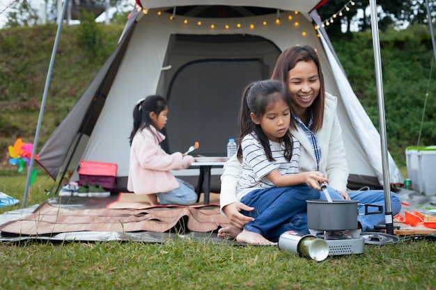 Photo cute asian child girl having fun to help her mother cooking outside the tent while camping with family.