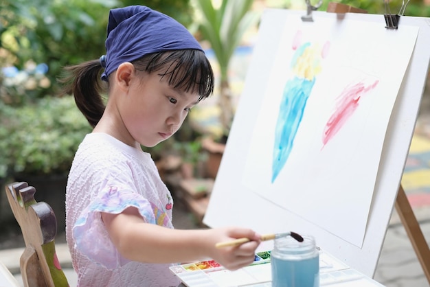 사진 귀여운 아시아 어린이 소녀는 페인트 브러쉬를 들고 종이에 그림을 그리고 선택적 초점을 맞추고 있습니다.