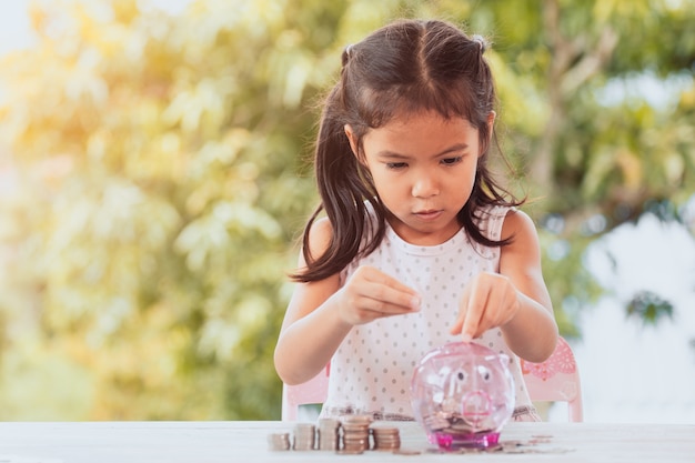 かわいいアジアの子供の女の子は、コインのスタックを作って、貯金箱にお金を入れて