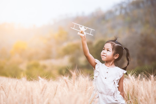 Cute asian child girl playing with toy wooden airplane in the barley field at sunset time
