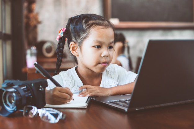 Cute asian child girl using laptop and writing on her notebook in the cafe