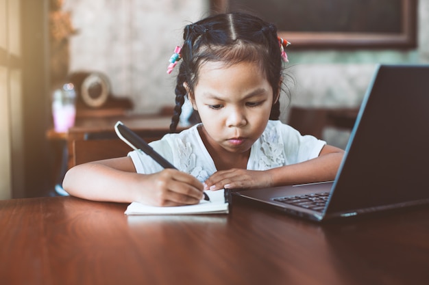 Cute asian child girl using laptop and writing on her notebook in the cafe