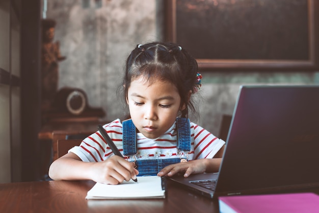 Cute asian child girl using laptop and writing on her notebook in the cafe