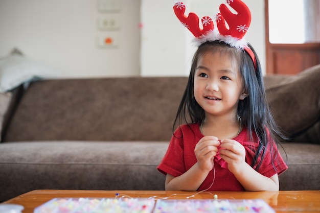 Cute asian child girl wearing christmas costume threading beads onto a string with intention and fun in home.