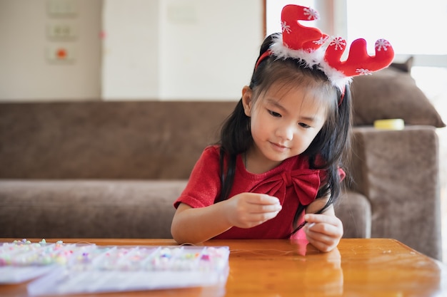 Cute asian child girl wearing christmas costume threading beads onto a string with intention and fun in home.