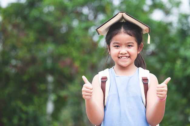 かわいいアジアの子供の女の子は、学校の袋と頭の上に本を置くと学校に行くのが大好き