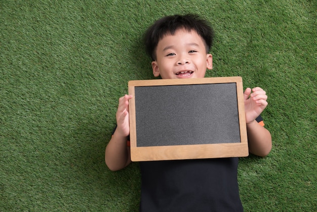 Bambino asiatico sveglio che si trova e che tiene lavagna su erba verde green