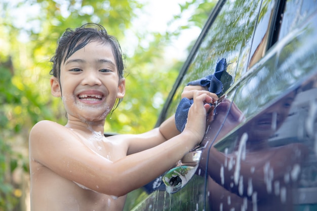 Cute asian child washing a car with hose on summer day