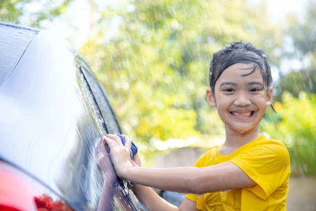 Cute asian child washing a car with hose on summer day