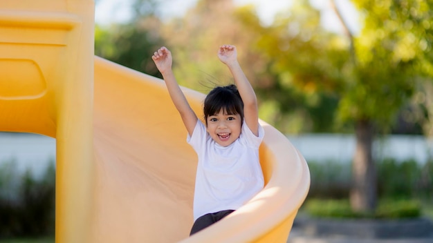 A cute Asian girl excitedly plays on a yellow playground slide