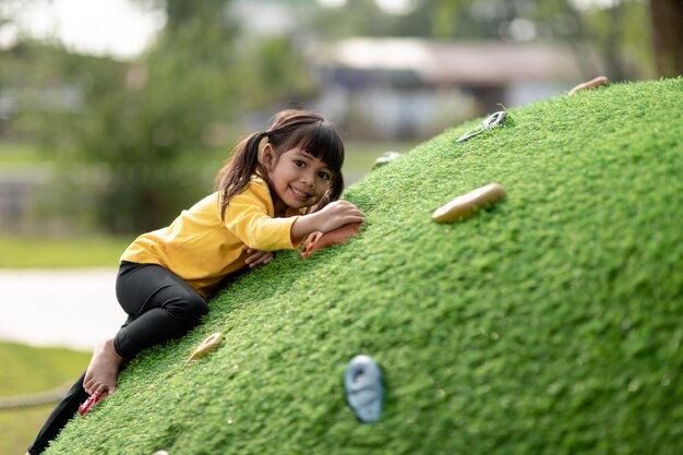 Photo cute asian girl having fun trying to climb on artificial boulders at schoolyard playground little girl climbing up the rock wall hand eye coordination skills development