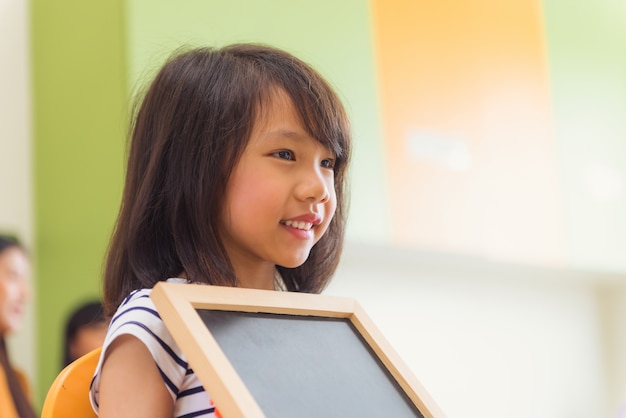 Cute asian girl holding blank blackboard with happy face in kindergarten classroom, kindergarten education concept, Vintage effect style pictures.