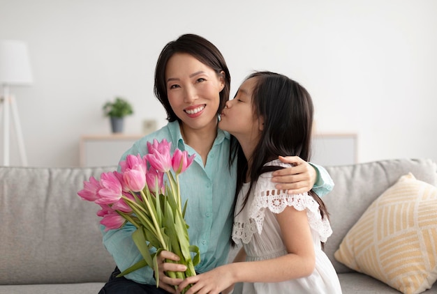 여성의 날 인사말을 위해 꽃을 주는 쾌활한 할머니와 키스하는 귀여운 아시아 소녀