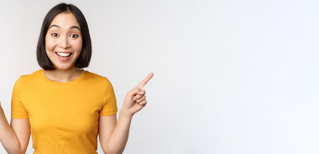 Cute asian girl pointing fingers sideways showing left and right promo two choices variants of products standing in yellow tshirt over white background