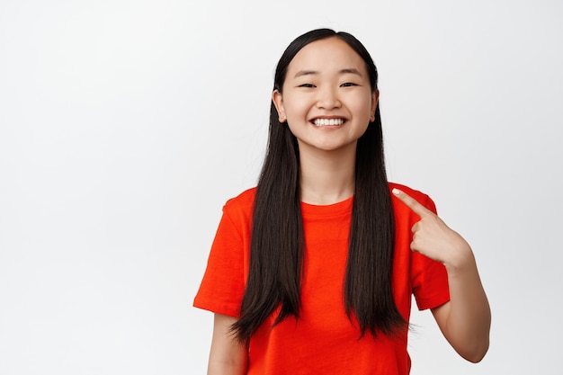 Cute asian girl points finger at herself, smiles and looks confident, pick me gesture standing on white.