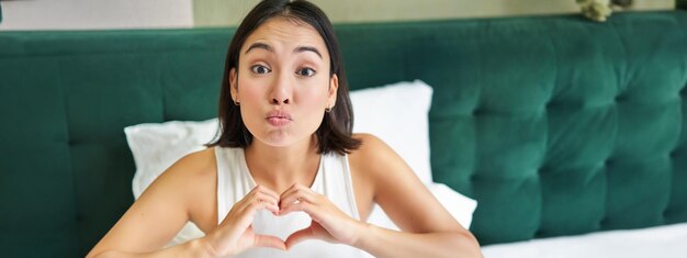 cute asian girl shows heart love sign lying in bed and smiling enjoying mornings lazy days in