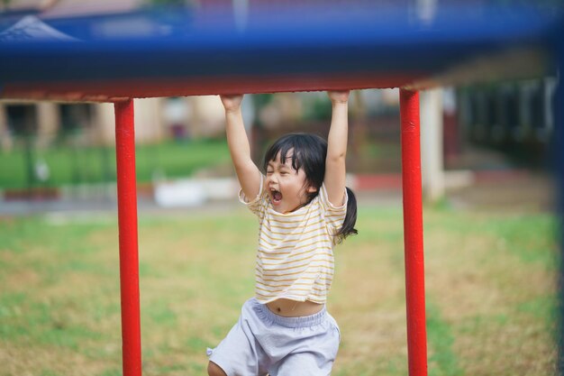 Photo cute asian girl smile play jumping on school kindergarten yard or playground healthy summer activity for children little asian girl climbing outdoors at playground child playing at playground