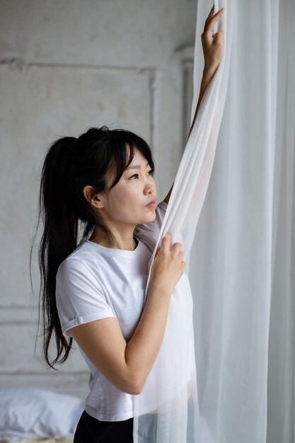 cute asian girl with dark long hair stands at the window and pulls back the white curtain