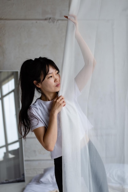 cute asian girl with dark long hair stands at the window and pulls back the white curtain