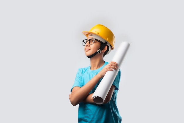 Cute Asian Indian happy kid wearing yellow construction helmet or safety hard hat, standing isolated on white wall holding blueprint